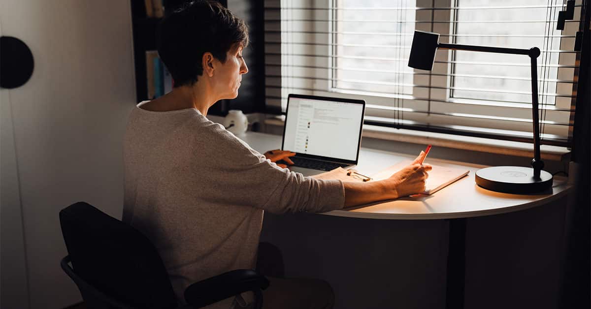 A woman working on laptop computer and writing down elevator pitch while sitting at table.